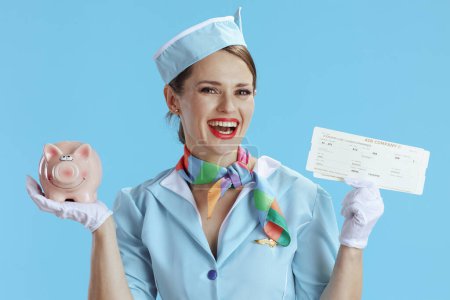 Photo for Happy elegant female stewardess against blue background in blue uniform with flight tickets and piggy bank. - Royalty Free Image