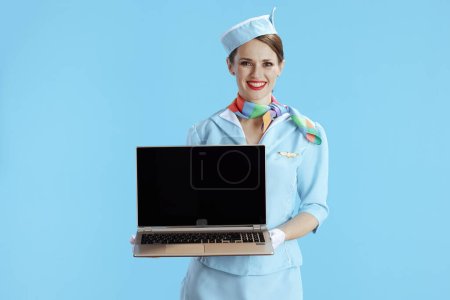 Téléchargez les photos : Élégante hôtesse de l'air souriante sur fond bleu uniforme bleu montrant écran blanc ordinateur portable. - en image libre de droit