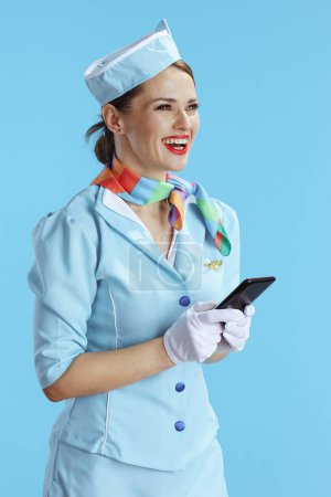 Photo for Happy modern female air hostess on blue background in blue uniform using smartphone. - Royalty Free Image