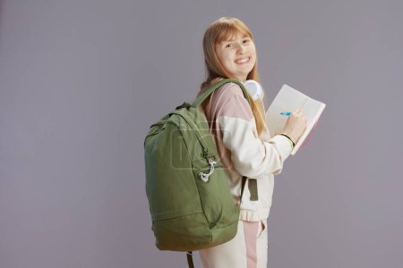 Photo for Portrait of smiling modern teenager girl in beige tracksuit with backpack, workbooks and headphones writing isolated on grey. - Royalty Free Image