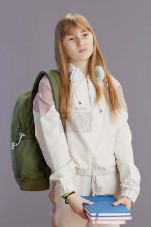 Photo for Sad modern teenager girl in beige tracksuit with backpack, workbooks and headphones isolated on grey background. - Royalty Free Image