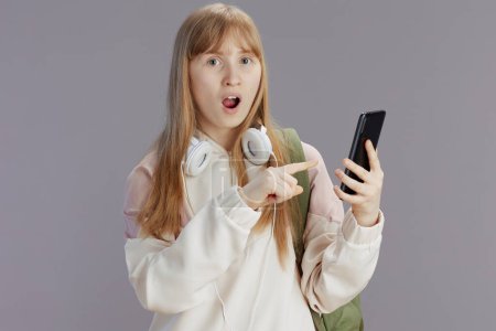 Foto de Sorprendida chica adolescente moderna en chándal beige con mochila y auriculares usando aplicación de teléfono inteligente aislado sobre fondo gris. - Imagen libre de derechos