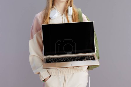 Photo for Closeup on girl in beige tracksuit with headphones showing laptop blank screen isolated on grey background. - Royalty Free Image
