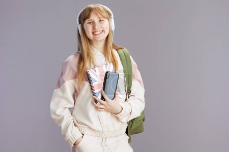 Photo for Portrait of happy trendy pupil in beige tracksuit with backpack, english flag workbook and headphones using smartphone isolated on grey background. - Royalty Free Image