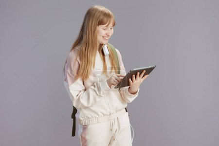 Foto de Sonriente chica adolescente de moda en chándal beige usando tableta PC contra gris. - Imagen libre de derechos