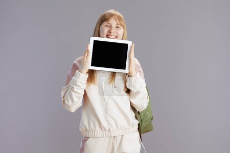Téléchargez les photos : Heureux élève à la mode en survêtement beige avec sac à dos montrant tablette PC écran vide isolé sur gris. - en image libre de droit