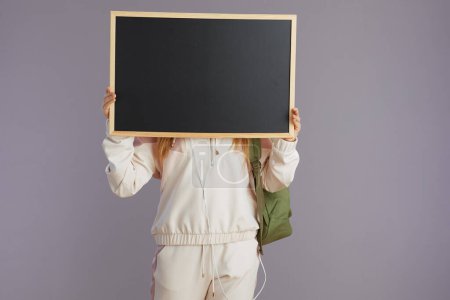 Photo for Trendy school girl in beige tracksuit with backpack showing blank board isolated on grey. - Royalty Free Image