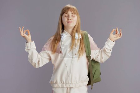 Photo for Relaxed trendy school girl in beige tracksuit with backpack and headphones isolated on grey. - Royalty Free Image