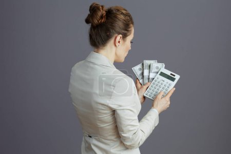 Photo for Seen from behind middle aged small business owner woman in a light business suit with calculator and dollars money packs isolated on gray. - Royalty Free Image