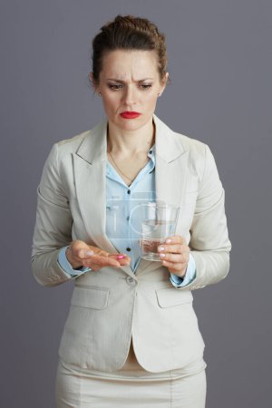 Foto de Cansada trabajadora elegante en un traje de negocios ligero con vaso de agua y píldora aislada en gris. - Imagen libre de derechos