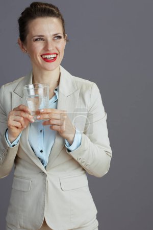 Téléchargez les photos : Heureuse femme d'affaires moderne dans un costume d'affaires léger avec verre d'eau isolé sur gris. - en image libre de droit