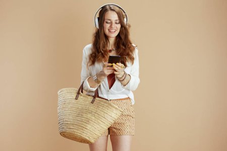 Photo for Beach vacation. happy modern middle aged housewife in white blouse and shorts isolated on beige with straw bag listening to the music with headphones and using smartphone. - Royalty Free Image