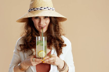 Téléchargez les photos : Vacances à la plage. femme moderne souriante en chemisier blanc et short isolé sur fond beige avec cocktail et chapeau de paille. - en image libre de droit