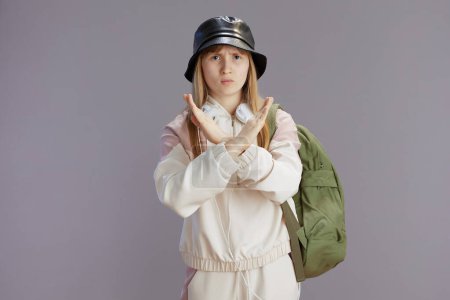 Photo for Portrait of unhappy modern girl in beige tracksuit with backpack, headphones and hat showing stop gesture isolated on grey background. - Royalty Free Image