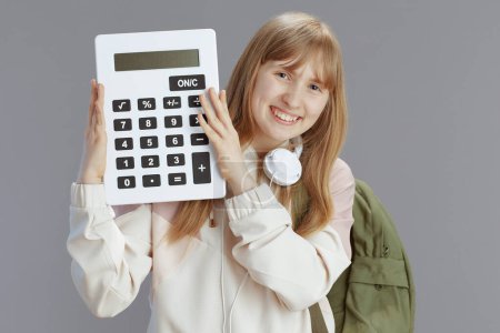 Photo for Portrait of smiling trendy teenager girl in beige tracksuit with backpack, calculator and headphones isolated on grey. - Royalty Free Image