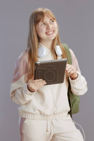 Photo for Happy trendy teenage girl in beige tracksuit with backpack, headphones and tablet PC against grey. - Royalty Free Image