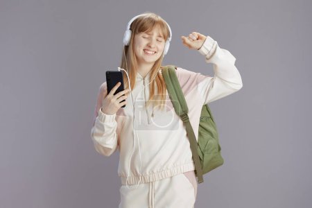 Téléchargez les photos : Écolière souriante et branchée en survêtement beige avec sac à dos dansant, écoutant la musique avec écouteurs et utilisant des applications smartphone isolées sur gris. - en image libre de droit