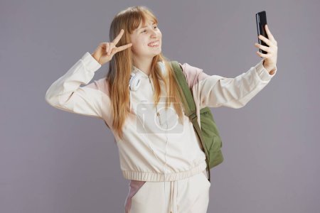 Foto de Sonriente chica de la escuela moderna en chándal beige con mochila y auriculares tomando selfie y el uso de teléfono inteligente contra gris mostrando gesto de victoria. - Imagen libre de derechos