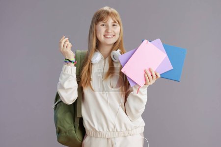Foto de Retrato de pupila de moda feliz en chándal beige con mochila, cuadernos de trabajo y auriculares aislados sobre dedos de fondo gris chasqueando. - Imagen libre de derechos