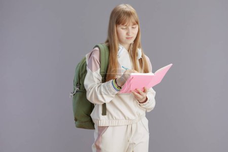 Photo for Modern teenage girl in beige tracksuit with backpack, workbooks and headphones writing against grey. - Royalty Free Image