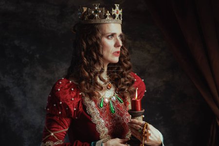 Téléchargez les photos : Reine médiévale en robe rouge avec bougie et couronne sur fond gris foncé. - en image libre de droit