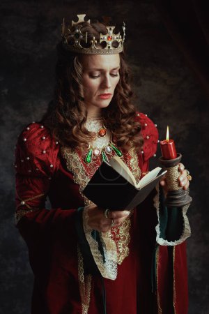 Foto de Reina medieval en vestido rojo con libro, vela y corona sobre fondo gris oscuro. - Imagen libre de derechos