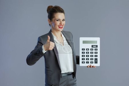 Téléchargez les photos : Heureux élégant 40 ans femme employée en costume gris avec calculatrice montrant pouces isolés sur gris. - en image libre de droit