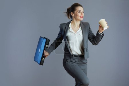 Photo for Carefree trendy 40 years old business woman in grey suit with coffee cup and briefcase walking isolated on gray background. - Royalty Free Image