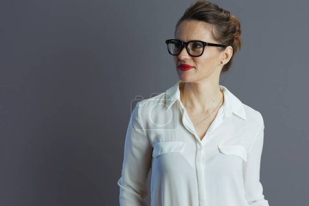 Téléchargez les photos : Coûteuse élégante femme d'âge moyen ouvrière en chemisier blanc avec des lunettes sur fond gris. - en image libre de droit