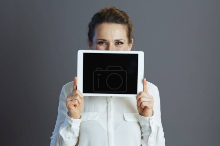 Photo for Trendy middle aged woman worker in white blouse showing tablet PC blank screen isolated on gray background. - Royalty Free Image