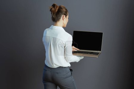 Photo for Seen from behind middle aged small business owner woman in white blouse with laptop against grey background. - Royalty Free Image
