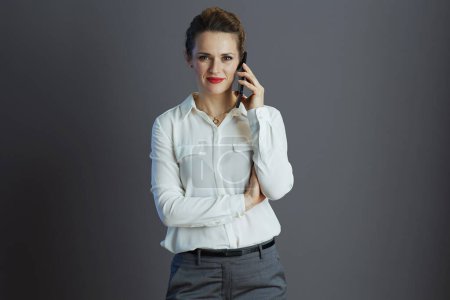 Téléchargez les photos : Femme d'âge moyen moderne employée en chemisier blanc parlant sur un téléphone mobile isolé sur fond gris. - en image libre de droit