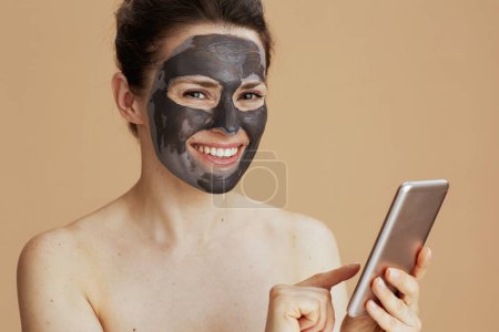 Téléchargez les photos : Portrait de jeune femme heureuse avec smartphone et masque facial isolé sur fond beige. - en image libre de droit
