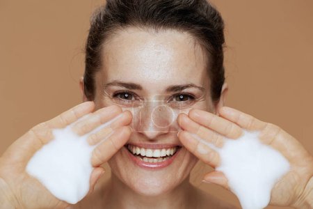Photo for Happy modern 40 years old woman with foaming facial cleanser washing face on beige background. - Royalty Free Image