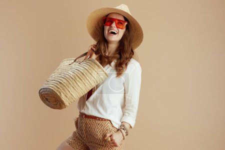 Photo for Beach vacation. happy trendy woman in white blouse and shorts isolated on beige with straw bag, summer hat and sunglasses. - Royalty Free Image