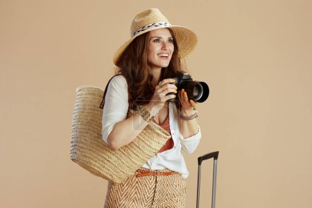 Photo for Beach vacation. happy trendy middle aged housewife in white blouse and shorts isolated on beige with straw bag, photo camera, trolley bag and straw hat. - Royalty Free Image