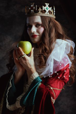 Photo for Medieval queen in red dress with green apple, white collar and crown on dark gray background. - Royalty Free Image