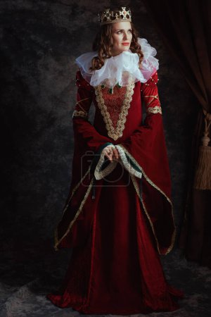 Full length portrait of pensive medieval queen in red dress with white collar and crown on dark gray background.