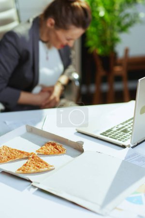 Foto de Lugar de trabajo sostenible. Primer plano sobre la mujer empleada en la oficina verde con pizza y portátil que tiene dolor de estómago. - Imagen libre de derechos