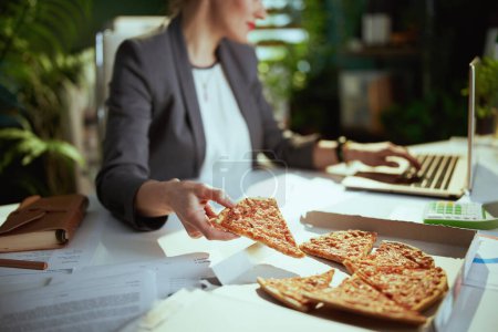 Photo for Sustainable workplace. Closeup on bookkeeper woman in a grey business suit in green office with pizza and laptop. - Royalty Free Image