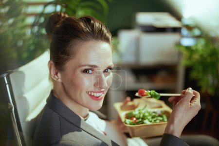 Foto de Lugar de trabajo sostenible. sonriente mujer de negocios moderna de mediana edad en un traje de negocios gris en la oficina verde moderna comer ensalada. - Imagen libre de derechos