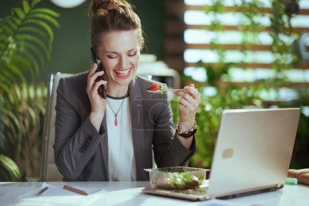 Foto de Lugar de trabajo sostenible. sonriente mujer de mediana edad moderna trabajadora en un traje de negocios gris en la oficina verde moderna con computadora portátil comer ensalada y hablar en un teléfono inteligente. - Imagen libre de derechos