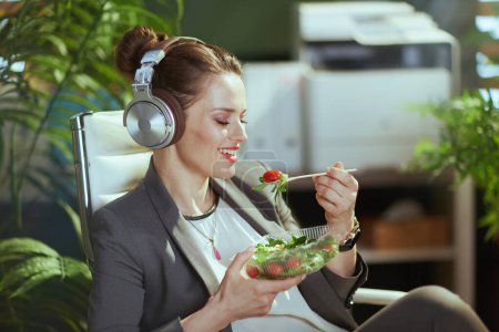 Téléchargez les photos : Un lieu de travail durable. femme propriétaire de petite entreprise moderne souriante dans un costume d'affaires gris dans un bureau vert moderne avec écouteurs manger de la salade. - en image libre de droit