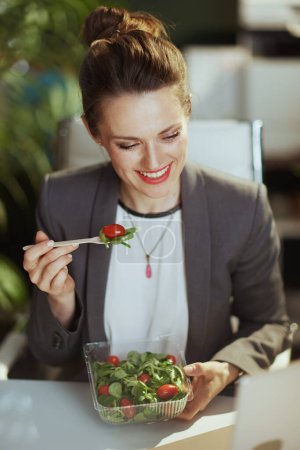 Foto de Lugar de trabajo sostenible. sonriente mujer contable moderna de mediana edad en un traje de negocios gris en una moderna oficina verde comiendo ensalada. - Imagen libre de derechos