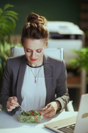 Téléchargez les photos : Un lieu de travail durable. travailleur femme d'âge moyen moderne dans un costume d'affaires gris dans un bureau vert moderne avec ordinateur portable manger salade. - en image libre de droit