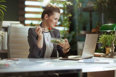 Téléchargez les photos : Un lieu de travail durable. heureuse femme comptable moderne de 40 ans dans un costume d'affaires gris dans un bureau vert moderne avec salade mangeant ordinateur portable. - en image libre de droit