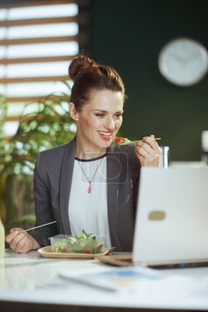 Foto de Lugar de trabajo sostenible. sonriente mujer de mediana edad moderna trabajadora en un traje de negocios gris en la oficina verde moderna con portátil comer ensalada. - Imagen libre de derechos