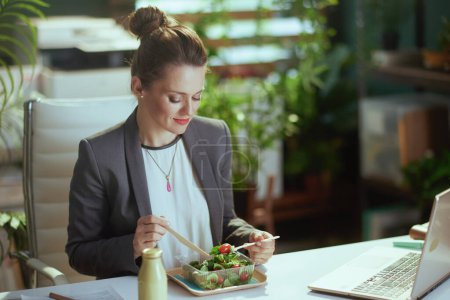 Foto de Lugar de trabajo sostenible. mujer contable moderna de mediana edad en un traje de negocios gris en la oficina verde moderna con portátil comer ensalada. - Imagen libre de derechos
