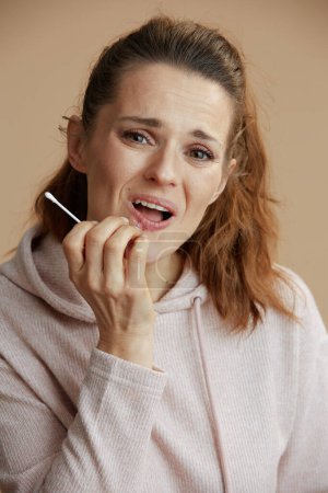 Téléchargez les photos : Femme d'âge moyen avec herpès sur les lèvres application de pommade en utilisant coton-tige sur fond beige. - en image libre de droit