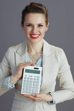 Photo for Smiling modern middle aged woman worker in a light business suit with calculator against gray background. - Royalty Free Image